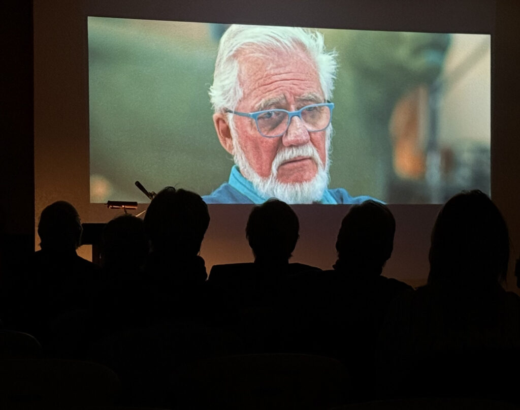 Audience watching a man on a large screen.